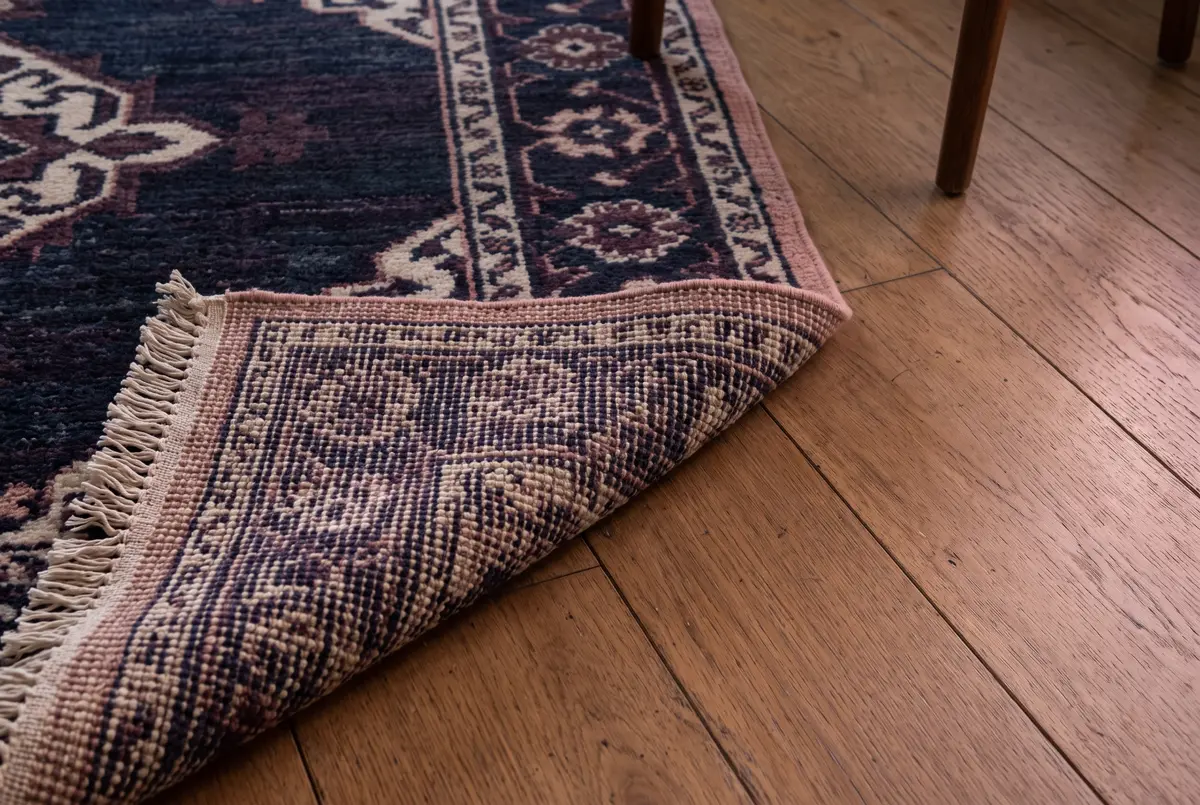 Illustration of a wool area rug with one corner turned over on a wood floor, showing its knotted back and a cream fringe.