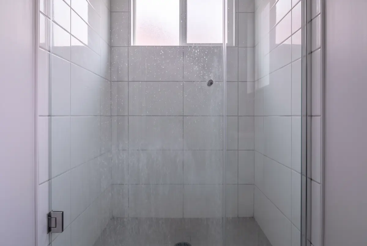 Illustration of a glass shower door and white tile behind it, both dotted with dried white mineral spots, lit from a small window.