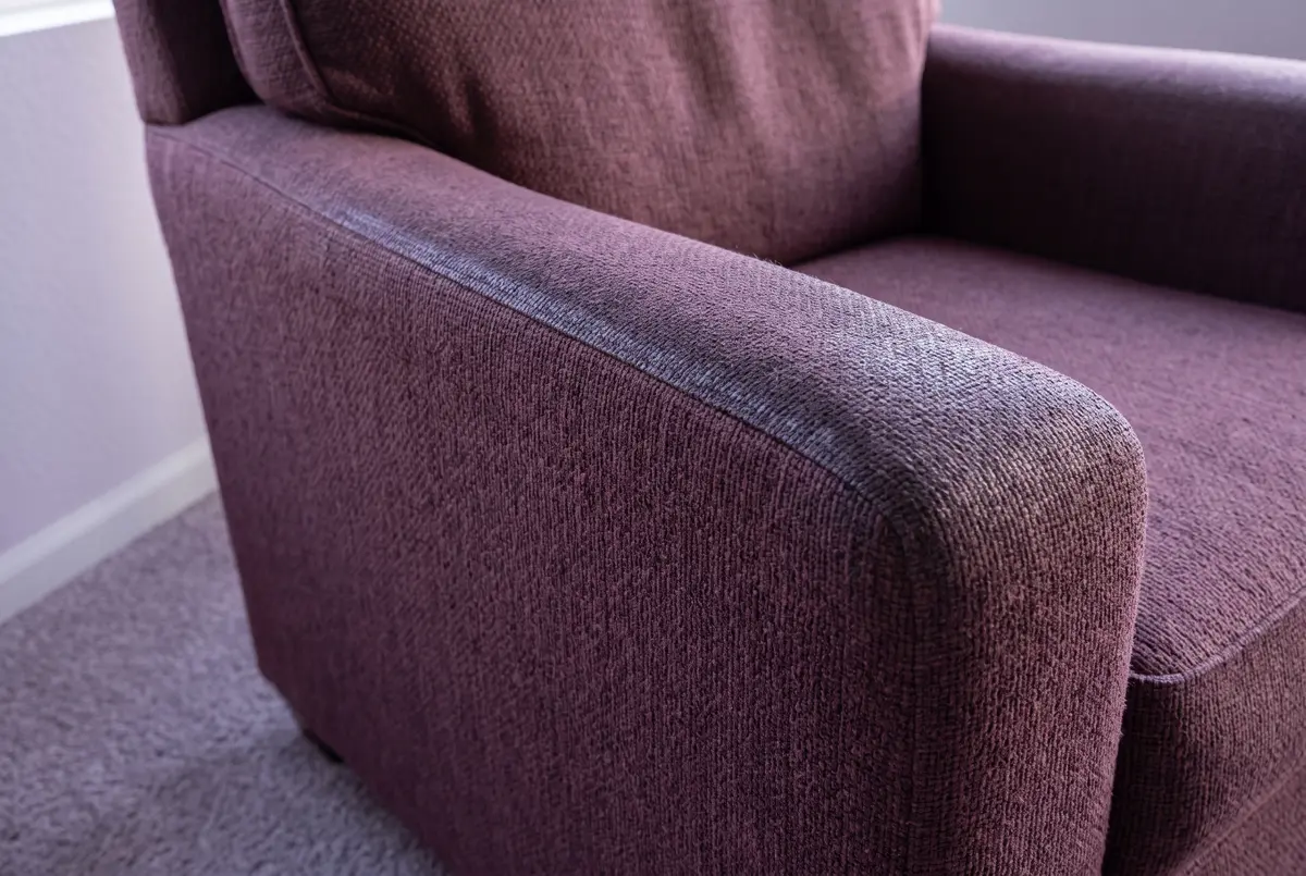 Illustration of the arm of a plum woven-fabric armchair, the weave worn shiny along the top where hands rest, with heather-gray carpet below.
